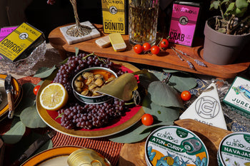 Elaborate spread of tinned fish, lemons, vegetables, tomatoes and decorative elements like glasses of wine and leaves