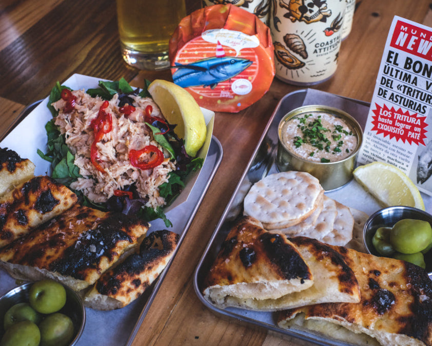 Plated food with bread, salad, and dips on a wooden table with a beer and newspaper.